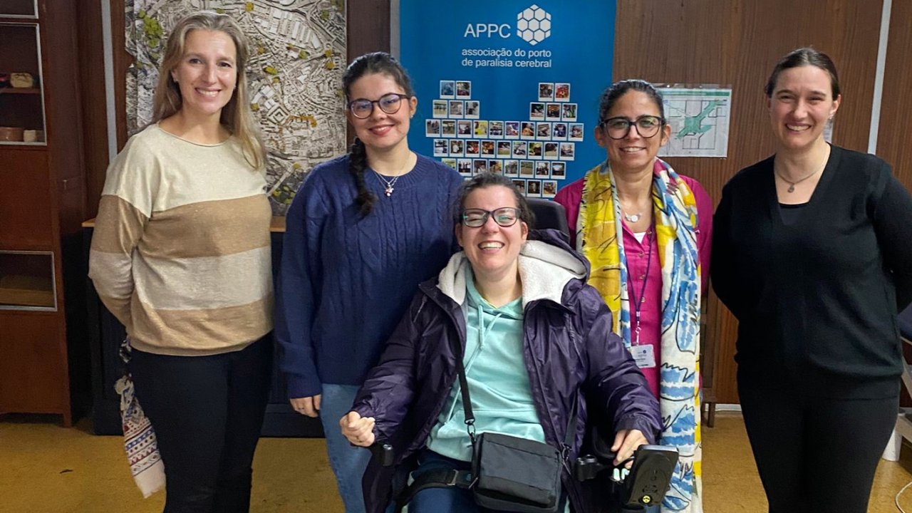 Grupo de cinco pessoas em espaço interior. Atrás do grupo um mapa afixado na parede e um cartaz (roll-up) de divulgação da Associação do Porto de Paralisia Cerebral.