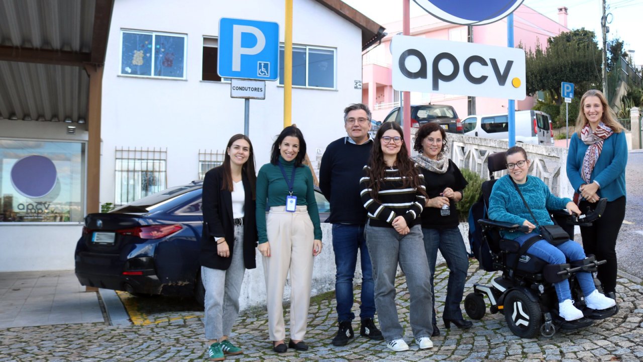 Grupo de sete pessoas junto à entrada de um edifício. Atrás do grupo está uma viatura estacionada. Vê-se ainda um sinal de trânsito e parte do logótipo da Associação de Paralisia Cerebral de Viseu (APCV).