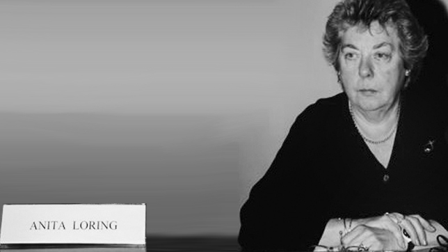 Fotografia, em tons cinza, de uma pessoa sentada junto de uma mesa e com as mãos sobrepostas. à esquerda uma placa com o nome Anita Loring.