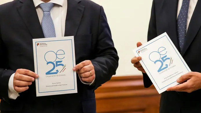 Fotografia parcial de duas pessoas, ambas com fato e gravata, que seguram nas mãos uma folha com indicação OE25.